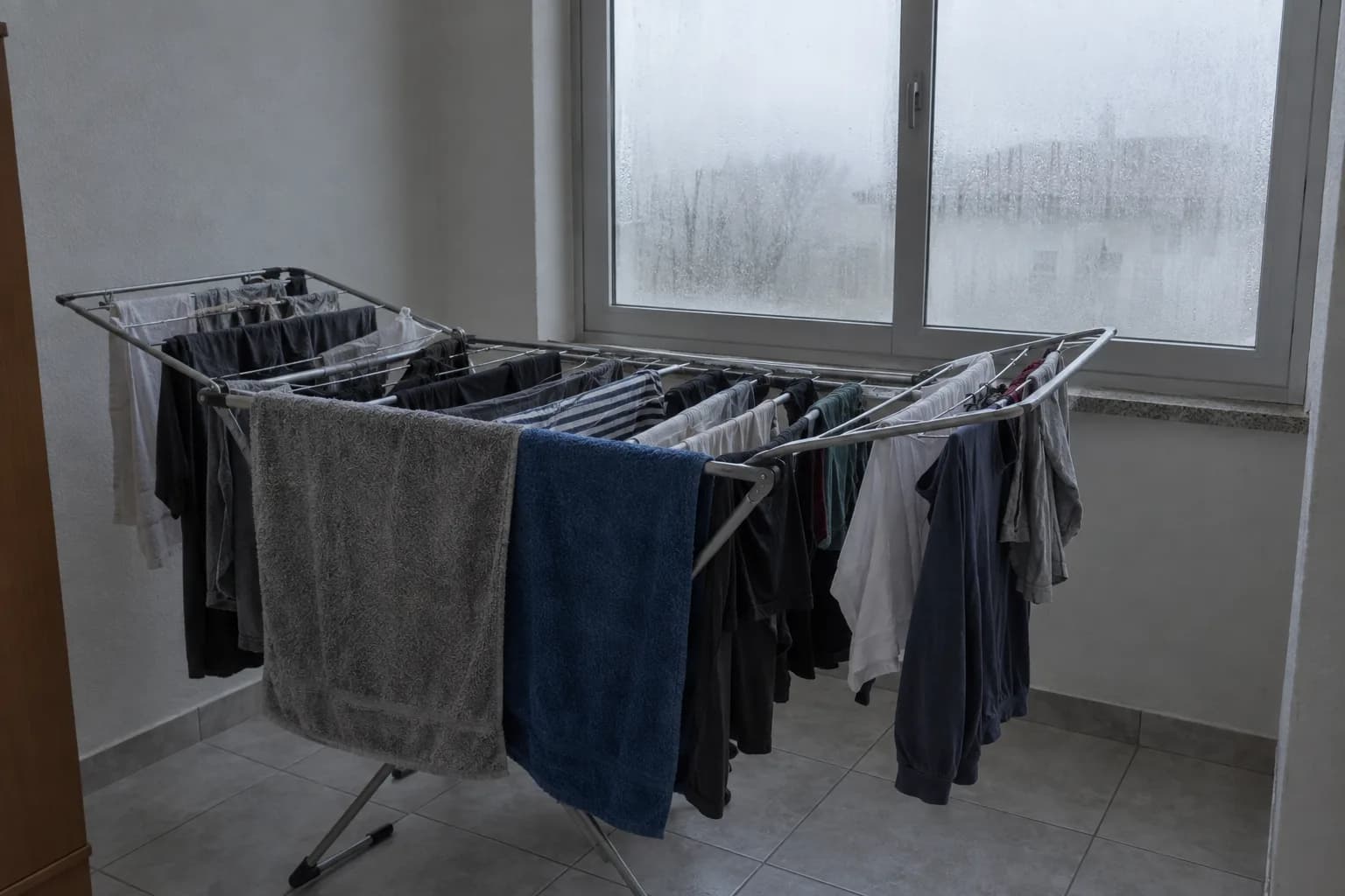 Divisão de um apartamento com um estendal de metal cheio de toalhas e roupa molhada junto a uma janela fechada, com o vidro embaciado e gotas de condensação, chão de ladrilho e paredes lisas
