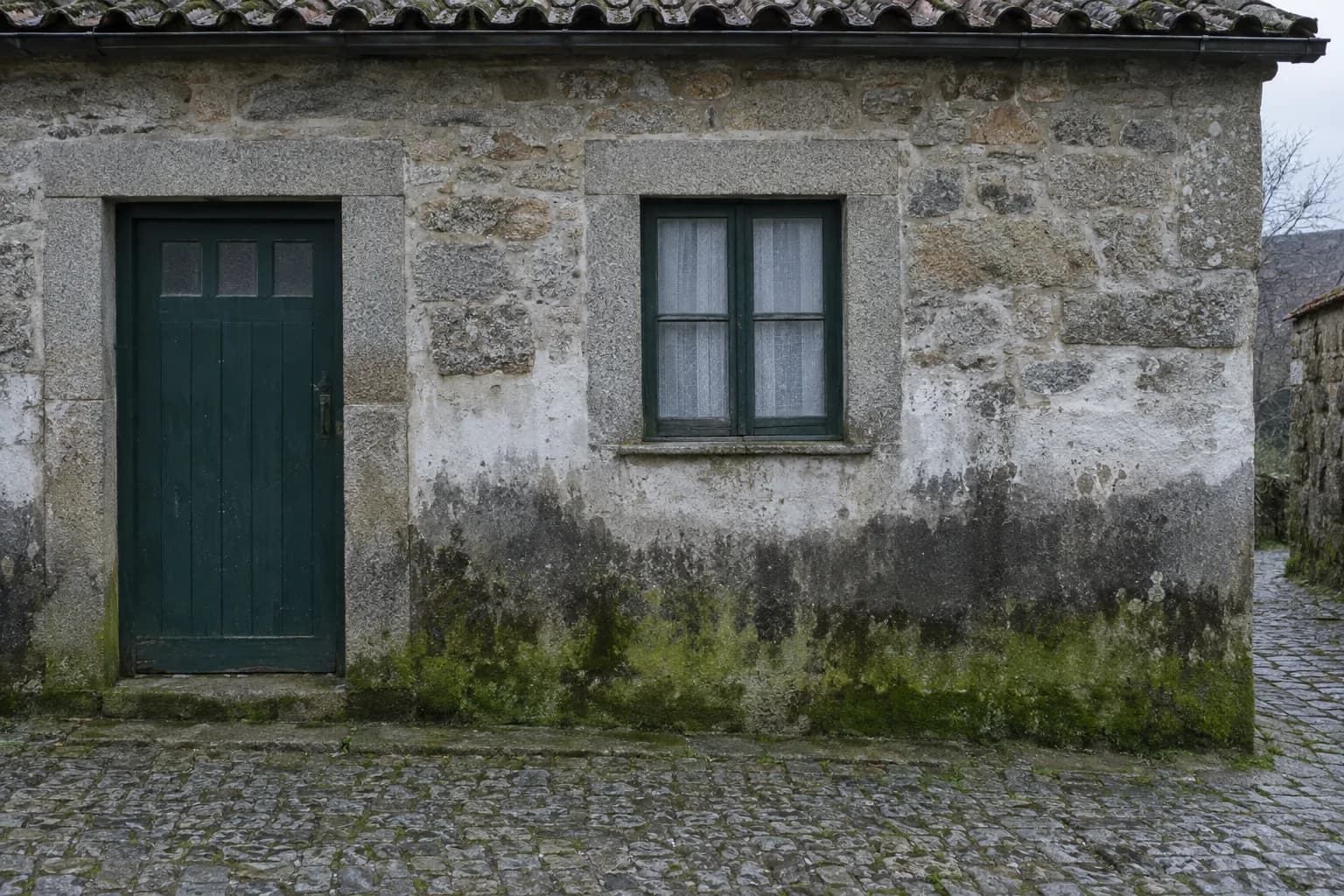 Parede exterior de uma casa de granito, com a base escurecida pela humidade e coberta de verdete, junto a um passeio de calçada