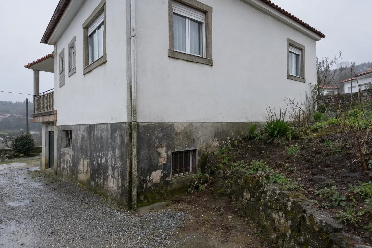 Lado de uma casa rebocada, com o terreno do jardim encostado à parede da cave, o reboco escurecido e a descascar na base e uma janela de cave com grade