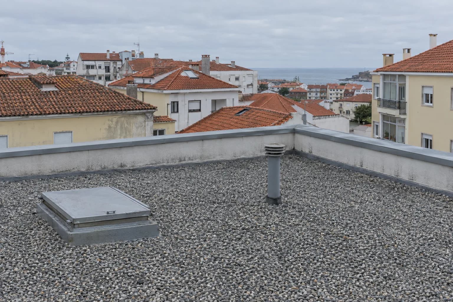 Cobertura plana de um prédio com gravilha, uma porta de acesso, um tubo de ventilação e a platibanda