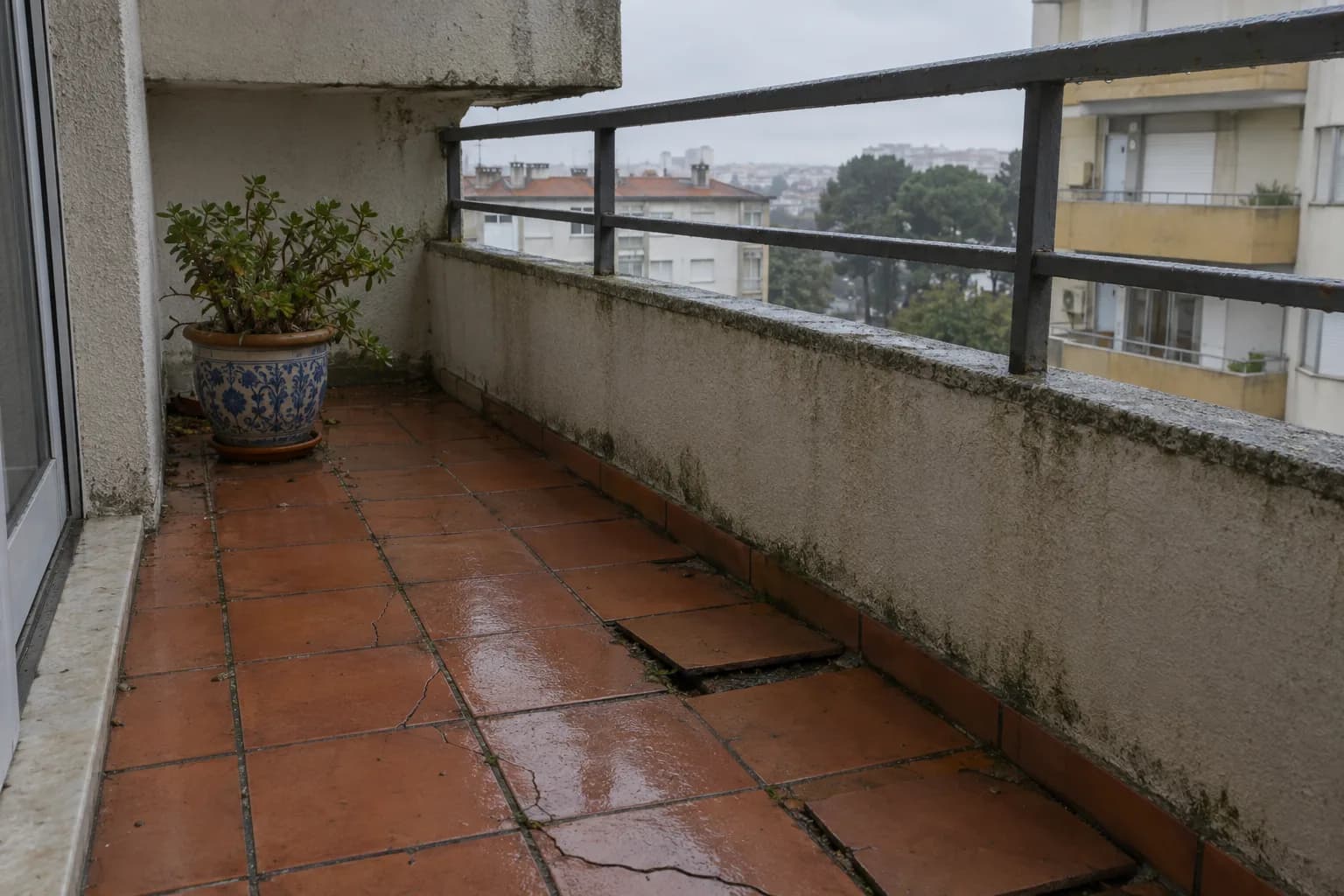 Varanda de um prédio dos anos 70, com ladrilho de barro molhado, peças fissuradas e soltas e a parede da varanda manchada junto ao chão