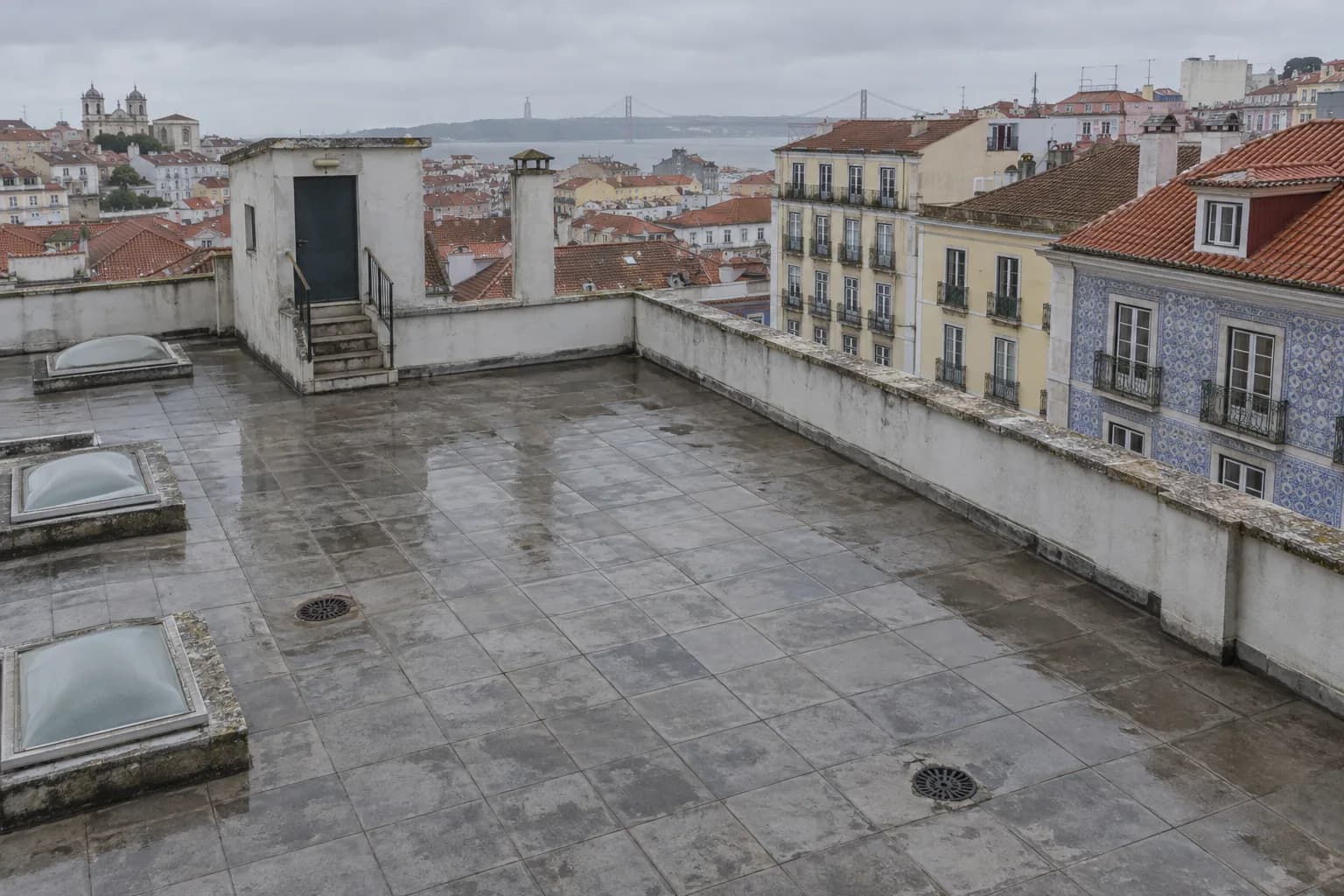 Vista de cima de um terraço grande de um prédio antigo em Lisboa, com ladrilho cinzento molhado, dois ralos, claraboias e uma platibanda baixa, e telhados da cidade ao fundo