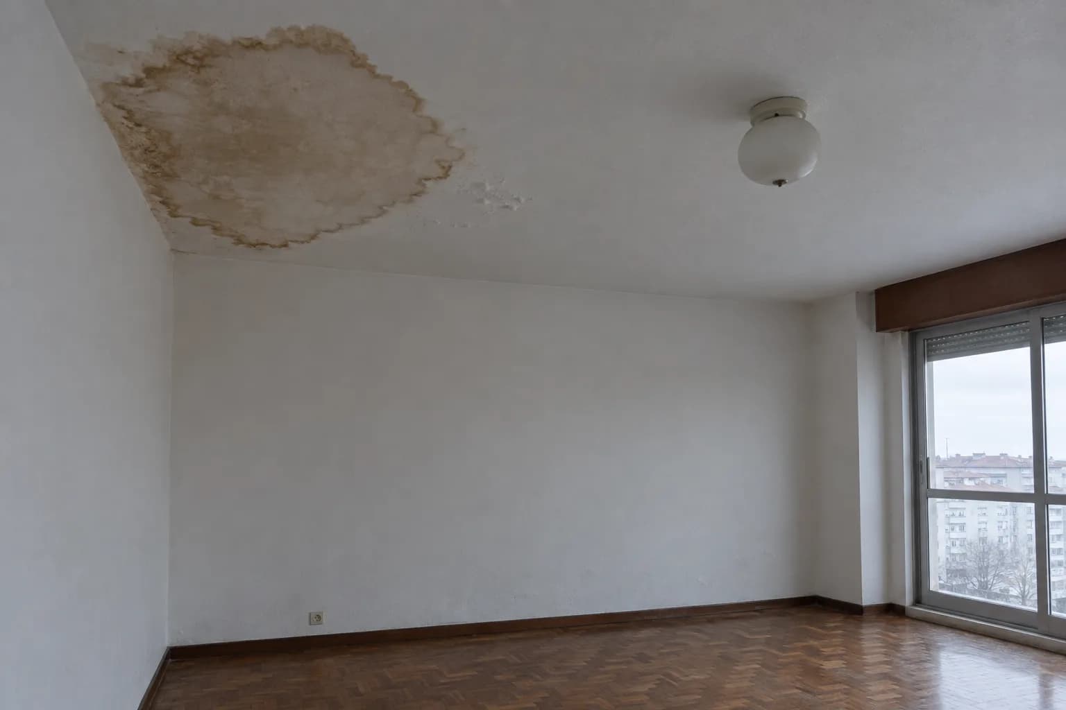 Teto de uma sala de um apartamento com uma grande mancha castanha de água junto ao canto, com a tinta a empolar, e uma janela com vista para prédios
